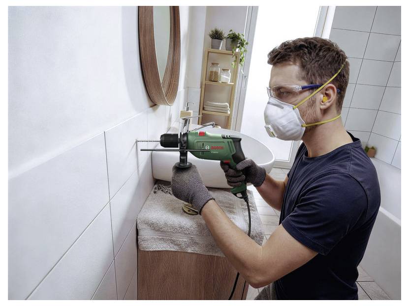 Ein Mann bohrt mit einer Elektrobohrmaschine ein Loch in eine Badezimmerwandkachel, während er Handschuhe, eine Schutzbrille und eine Maske trägt.