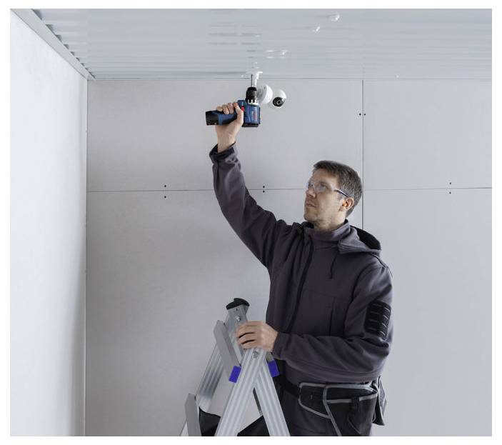 Ein Arbeiter auf einer Leiter benutzt einen Elektroschrauber über Kopfhöhe, um ein Deckenpanel in einem Raum unter Konstruktion zu installieren oder zu befestigen.