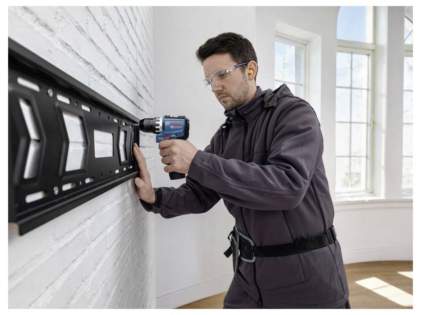 Ein Mann verwendet einen Elektrobohrer, um ein schwarzes Wandpaneel an einer weißen Ziegelmauer im Innenbereich in der Nähe von hellen Fenstern anzubringen.