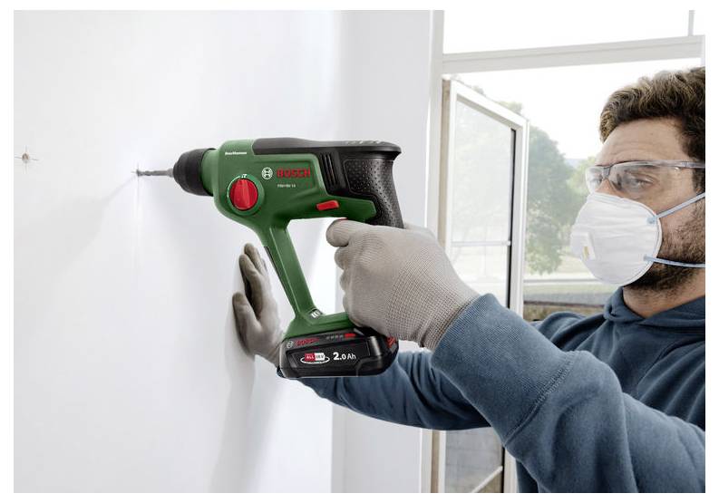Ein Mann, der eine Schutzbrille, eine Maske und Handschuhe trägt, bohrt mit einem Bosch-Akkubohrer in eine weiße Wand neben einem offenen Fenster.