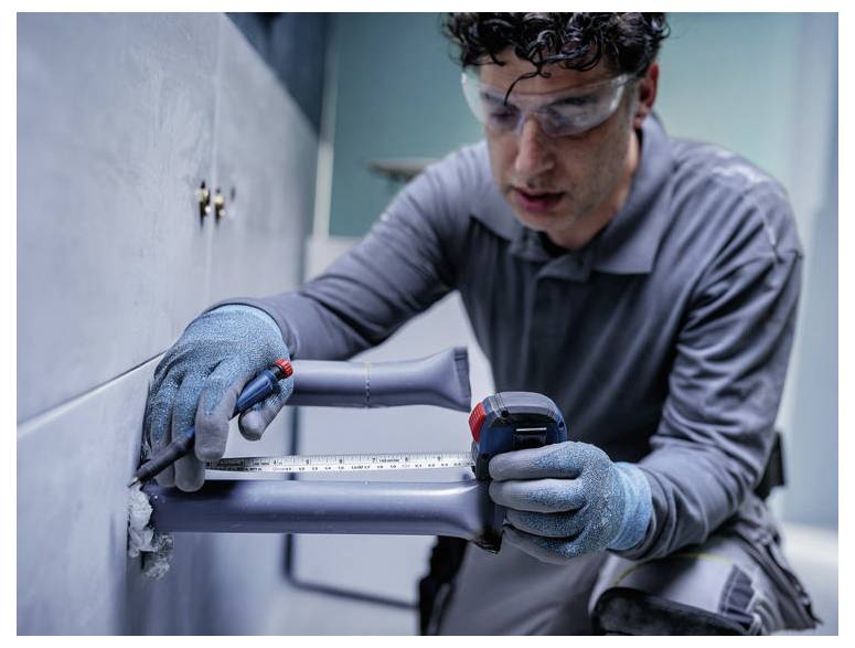 Techniker mit Handschuhen und Schutzbrille installiert ein Metallrohr unter einem Waschbecken mit einem Schraubenschlüssel und einem Messstab.