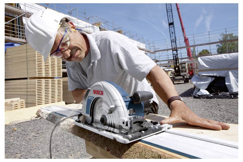 Bauarbeiter mit Schutzhelm und Schutzbrille schneidet ein Brett mit einer Bosch-Kreissäge auf einer Außenbaustelle.