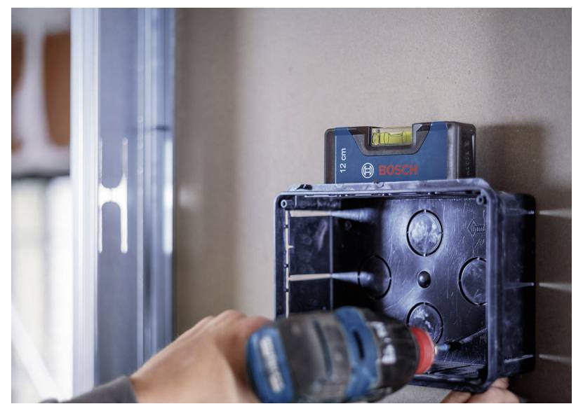 Eine Person bohrt mit einer Bosch-Elektrobohrmaschine ein Loch in eine wandmontierte Elektrobox und verwendet eine Wasserwaage, um sie ausgerichtet zu halten.