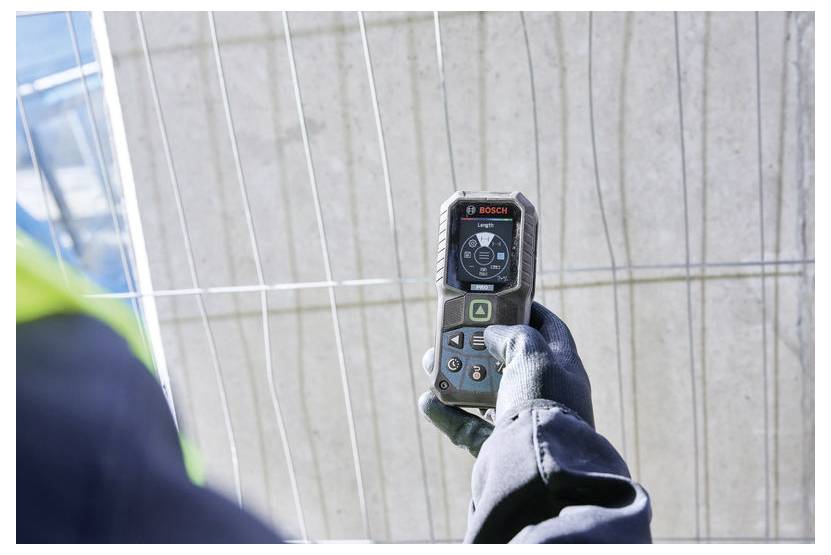 Ein Arbeiter mit Handschuhen hält ein tragbares Bosch-Laser-Entfernungsmessgerät gegen eine Wand und misst eine vertikale Innenfläche.