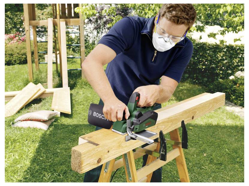 Ein Mann mit Schutzbrille und Maske schneidet einen Holzbalken mit einer Bosch-Kreissäge auf Sägeböcken im Freien.