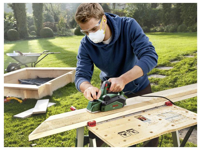 Ein Mann mit einer Maske und Schutzbrille hobelt ein Holzbrett im Freien auf einer Werkbank, mit Werkzeugen und Holz in der Nähe.