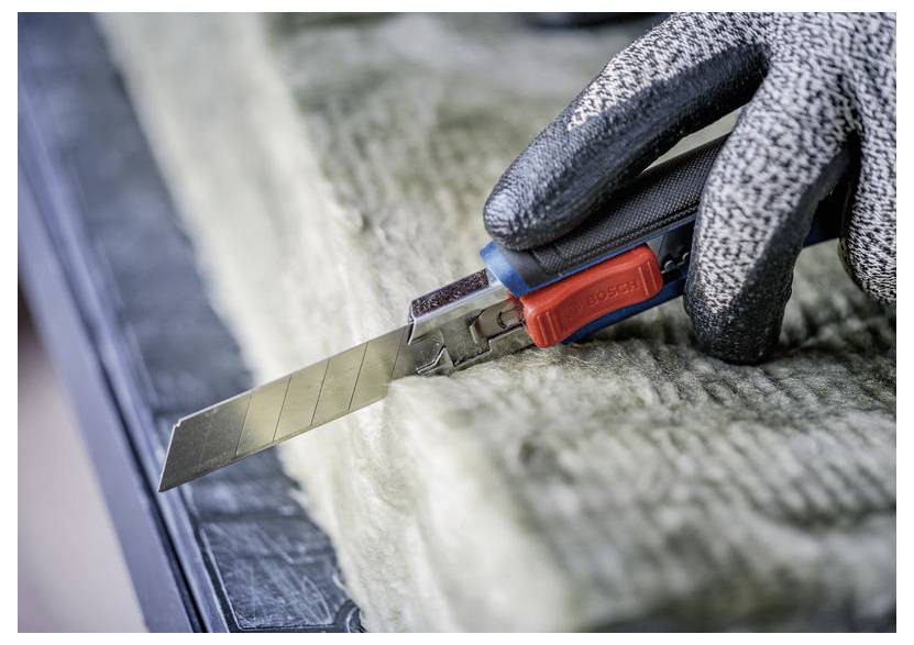 Handschuh tragende Hand benutzt ein Universalmesser, um dickes Isolationsmaterial auf einer Arbeitsfläche zu schneiden.