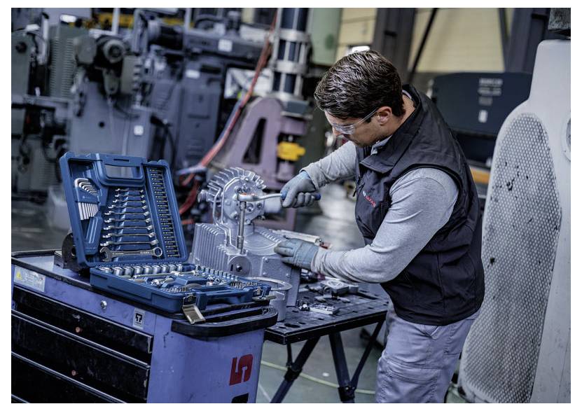 Ein Mechaniker mit Handschuhen und Schutzbrille benutzt einen Schraubenschlüssel zur Reparatur einer Maschine neben einem offenen blauen Steckschlüssel-Werkzeugsatz in einer Werkstatt.
