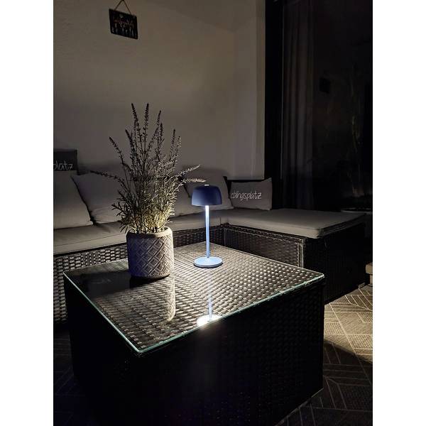 Gemütliche Terrassenlounge in der Nacht mit Rattanmöbeln, einem Glastisch, einer Topfpflanze und einer kleinen leuchtenden Tischleuchte.