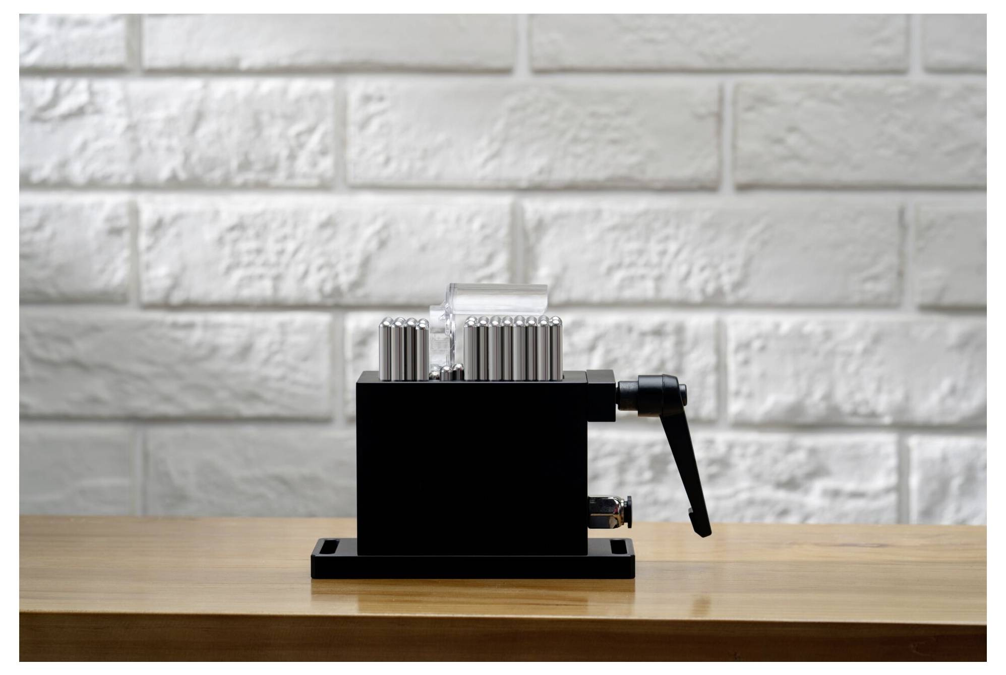 Schwarze manuelle Kaffeemühle mit einer Seitenkurbel auf einem Holztisch vor einer weißen Ziegelmauer.
