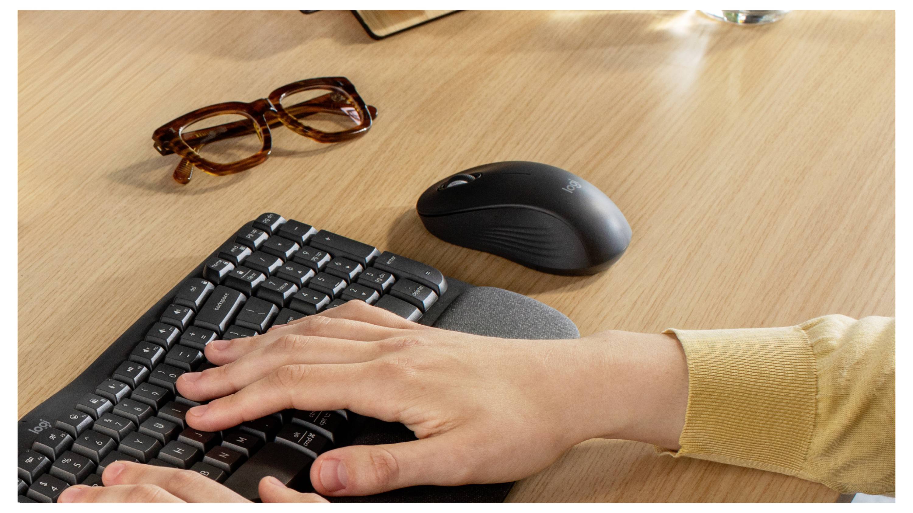 Hand tippt auf einer Tastatur an einem Schreibtisch mit einer Maus und Brille in der Nähe.