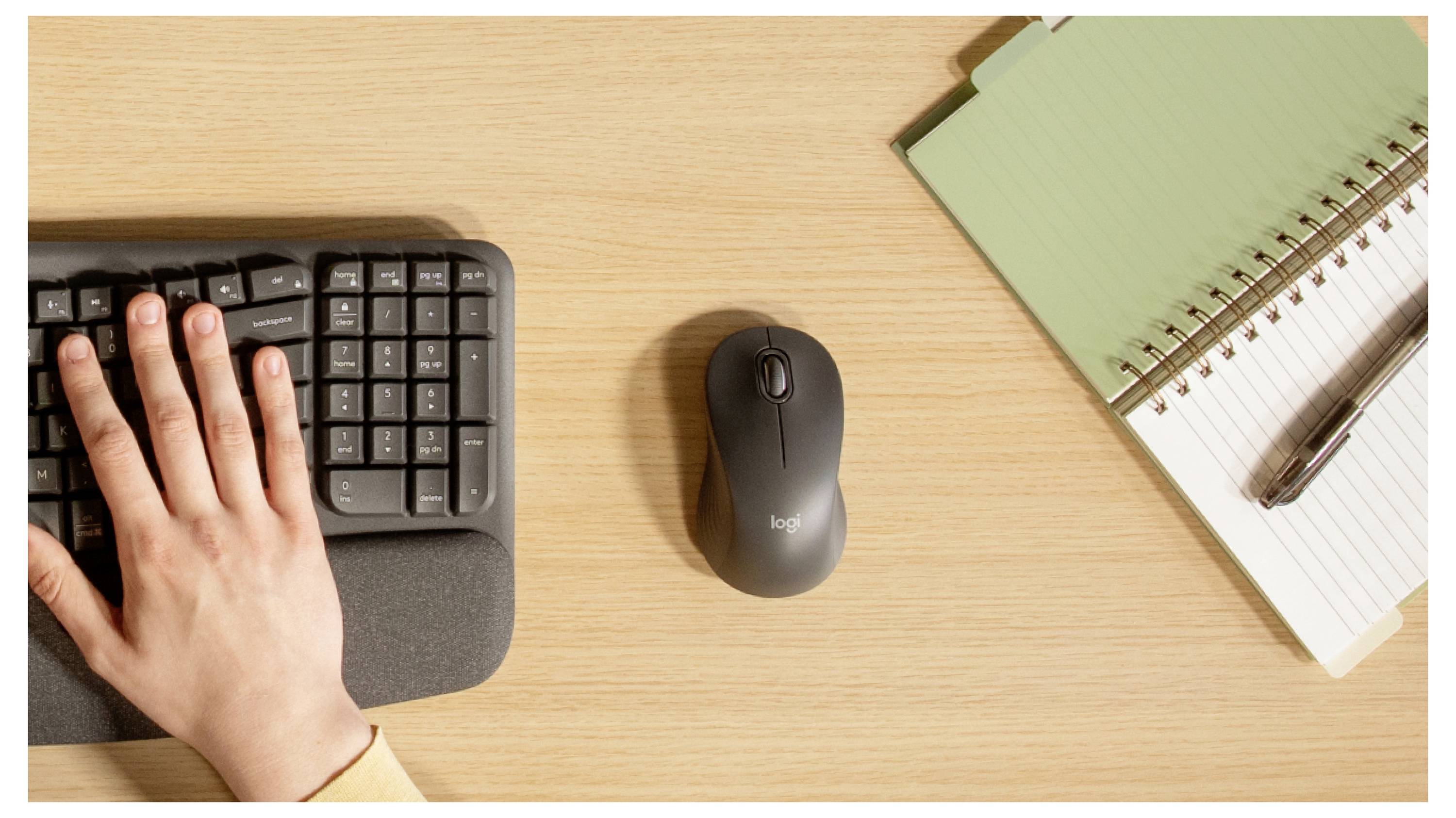 Draufsicht auf einen Schreibtisch mit einer Hand auf einer Tastatur, einer Maus in der Mitte und einem Notizbuch mit Stift auf der rechten Seite.