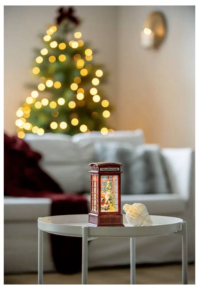 Eine leuchtende Weihnachtslaterne auf einem kleinen Tisch im Vordergrund, mit einem unscharfen Weihnachtsbaum und Sofa im Hintergrund.