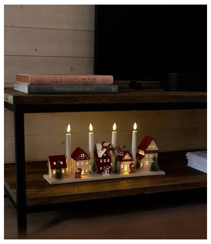 Beleuchtetes Miniatur-Weihnachtsdorf mit Kerzen auf einem Holzregal unter einem Fernseher, mit gestapelten Büchern oben und Papieren auf der rechten Seite.