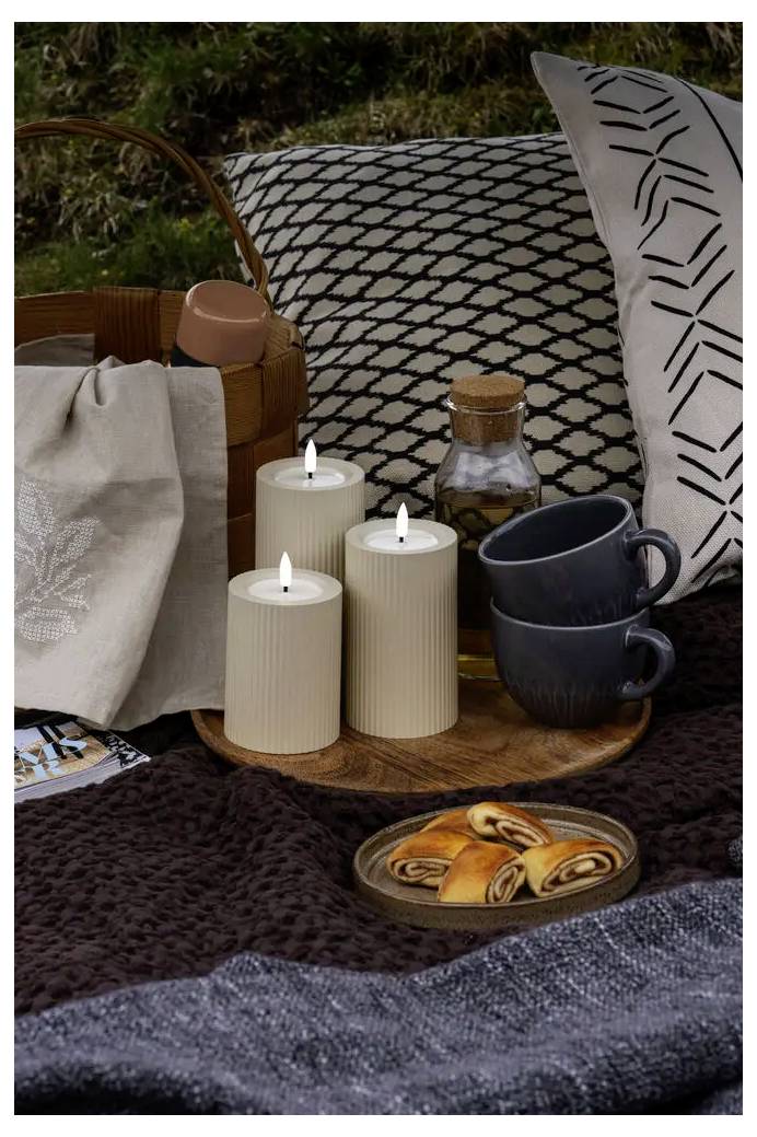 Picknick-Setup bei Kerzenlicht mit drei brennenden Säulenkerzen, gestapelten Bechern, einer Flasche und Gebäck auf einem Tablett neben einem Korb und gemusterten Kissen.