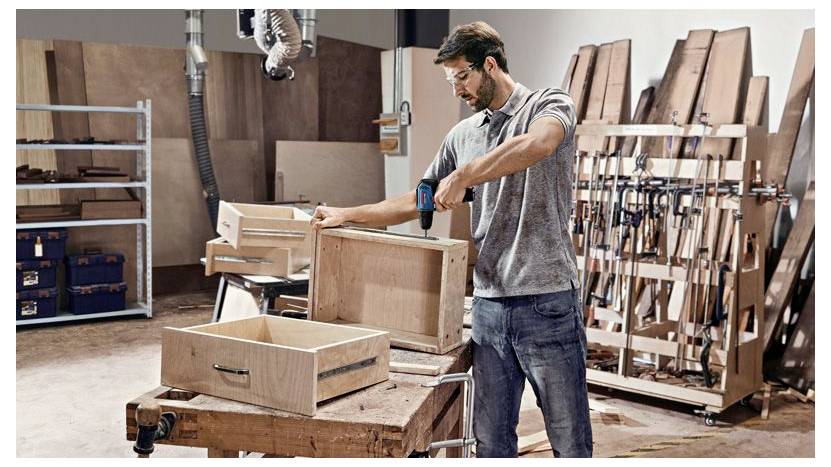 Ein Mann bohrt und montiert eine hölzerne Kiste in einer Werkstatt, umgeben von Werkzeugen und gestapeltem Holz.