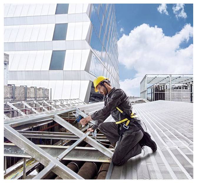 Bauarbeiter in einem gelben Schutzhelm kniet auf einem Stahlkonstruktionsrahmen eines Daches und arbeitet an einer erhöhten Gebäudestruktur vor einer modernen Glasfassade und dem Himmel.