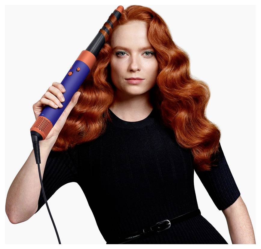 Porträt einer rothaarigen Frau, die ein blau-orange gefärbtes Lockenstab gegen ihren Kopf hält, vor einem schlichten weißen Hintergrund.