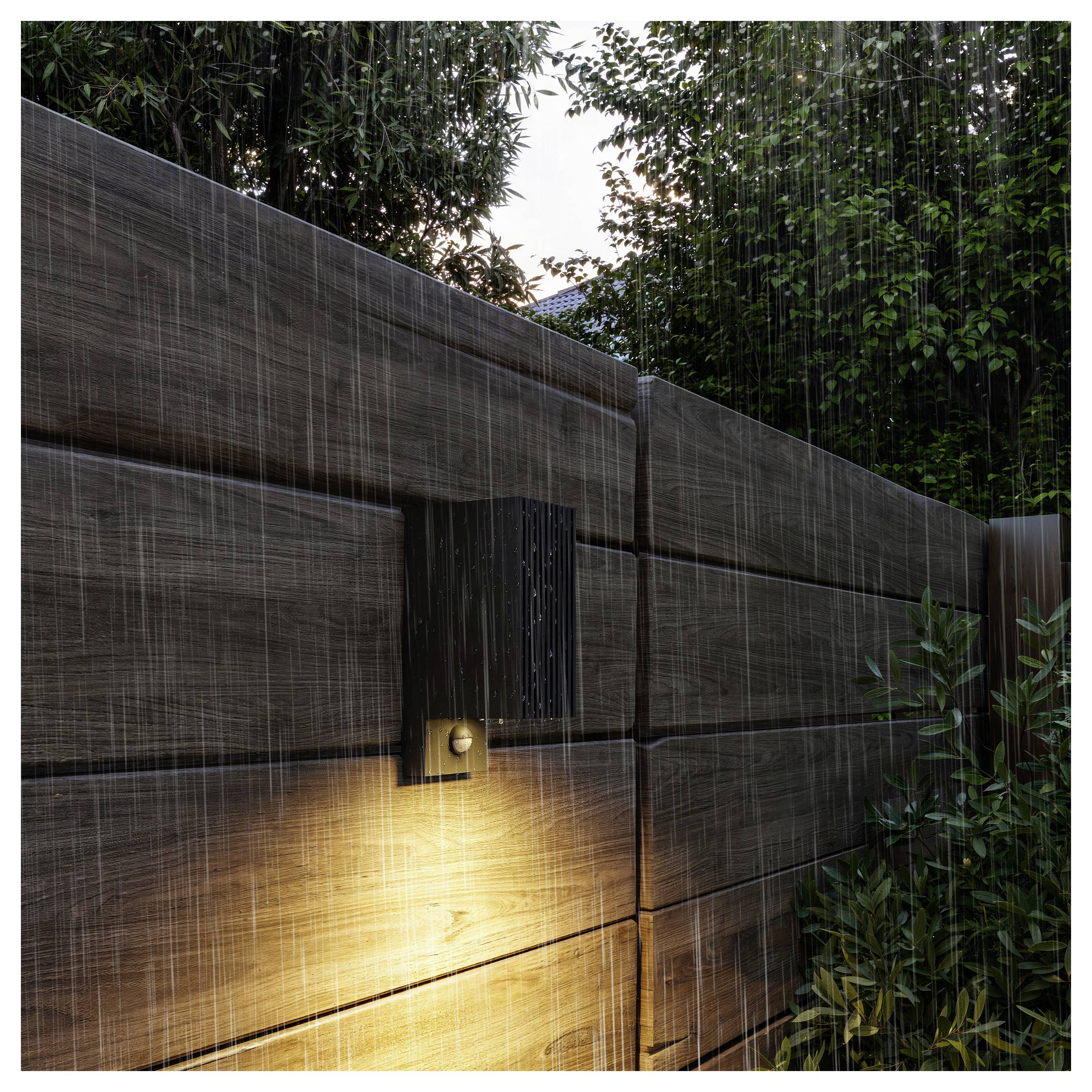Regen fällt auf ein modernes Holztor, das von einem warmen Wandlicht beleuchtet wird, mit dichtem Grün und einem Hausdach dahinter sichtbar.