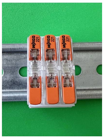 Drei orangefarbene WAGO 221-Drahtverbinder auf einer Metallhutschienenoberfläche vor grünem Hintergrund befestigt.