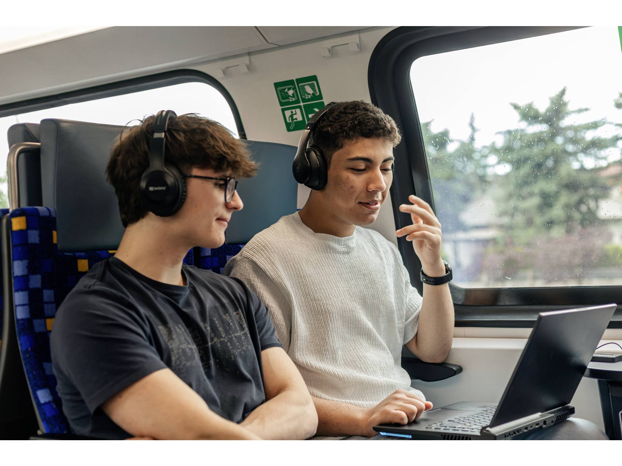Zwei junge Männer mit Kopfhörern sitzen in einem Zug, schauen auf einen Laptop und unterhalten sich leise am Fenster.