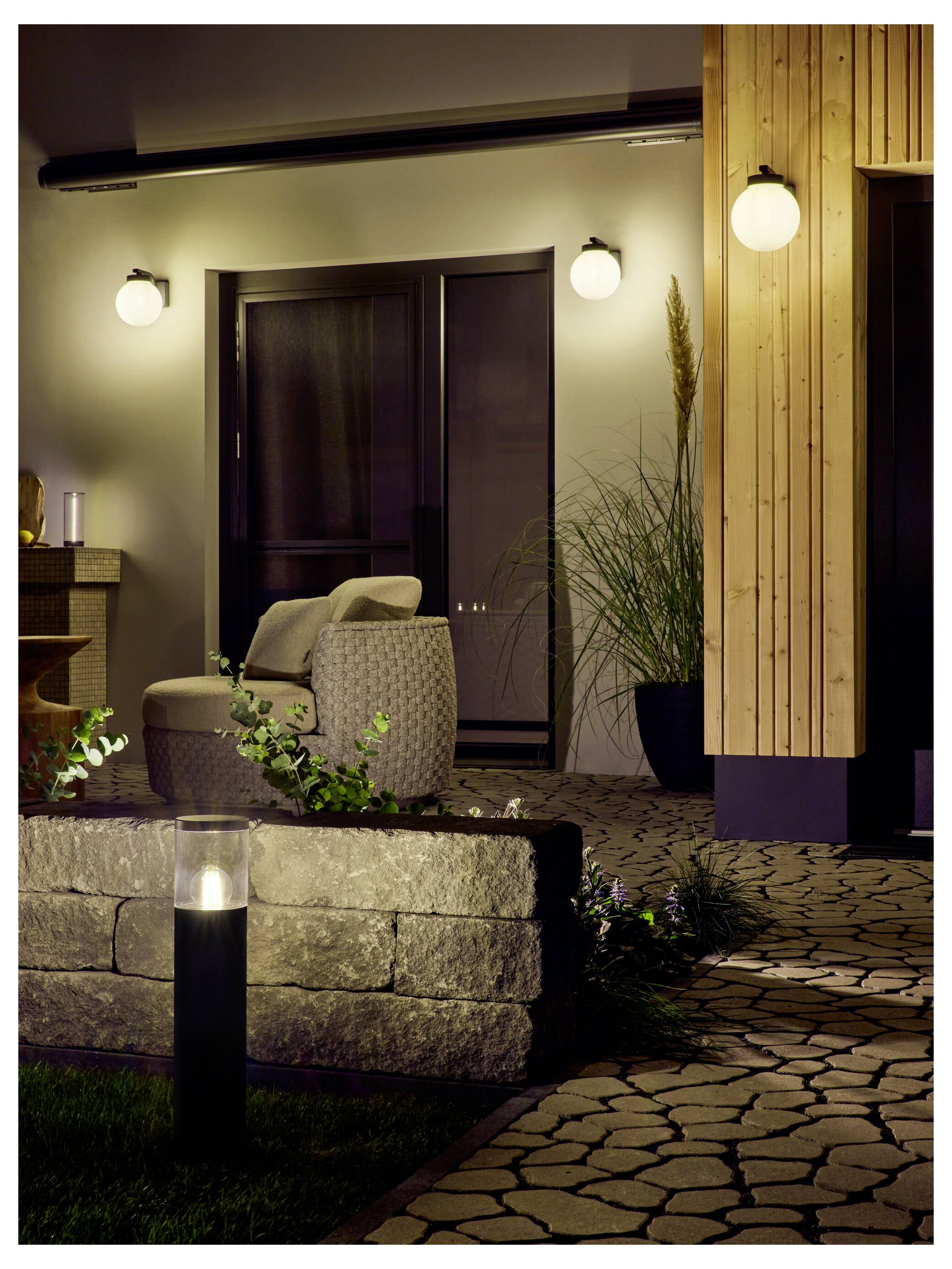 Attraktiver Hauseingang bei Nacht mit Wandleuchten, gepflastertem Gehweg, Topfpflanzen und einem Stuhl auf einer kleinen Terrasse.