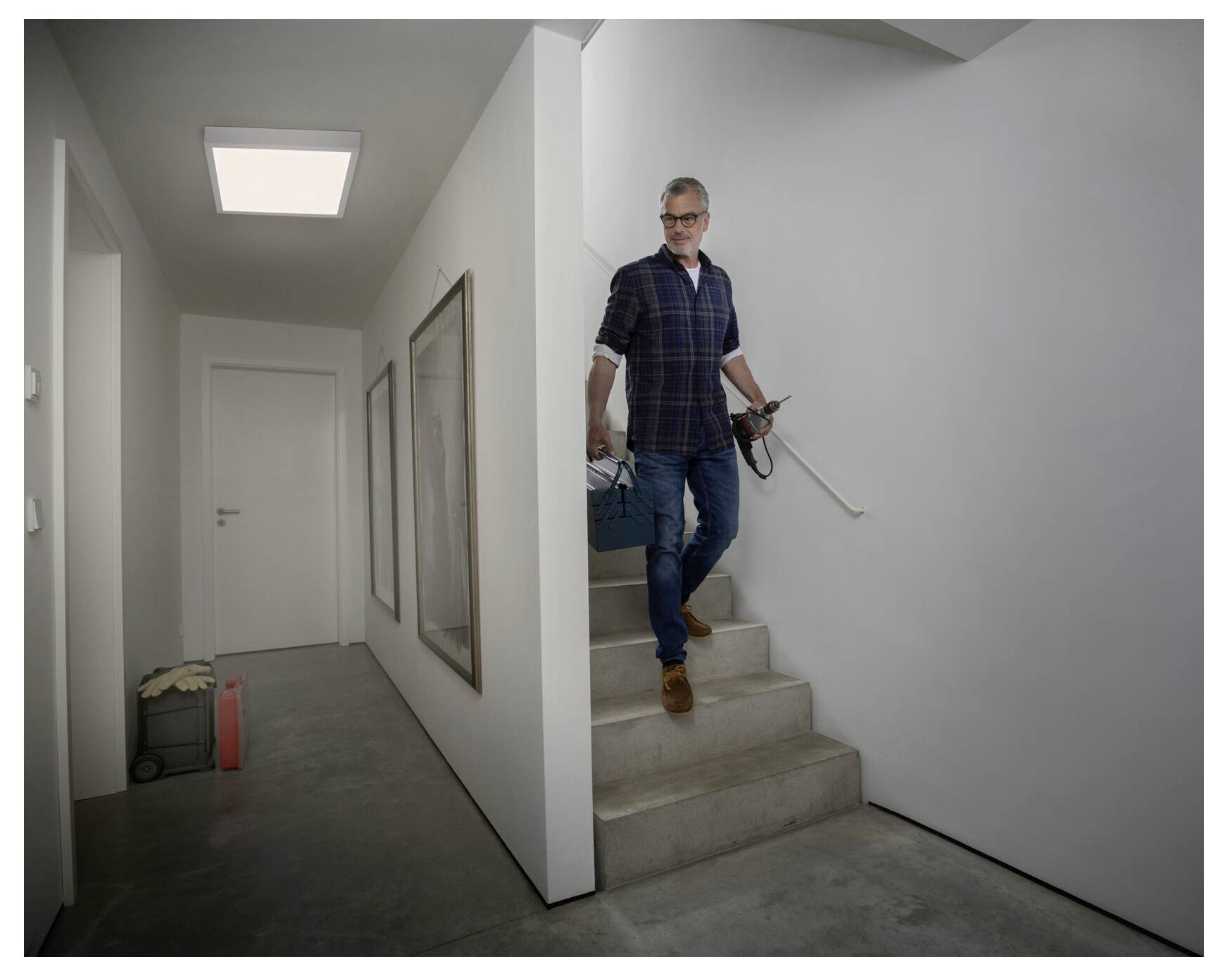 Ein Mann trägt Werkzeuge und eine Werkzeugkiste eine enge Treppe in einem modernen Flur hinunter, was auf Haushaltsreparaturen oder Renovierungsarbeiten hindeutet.