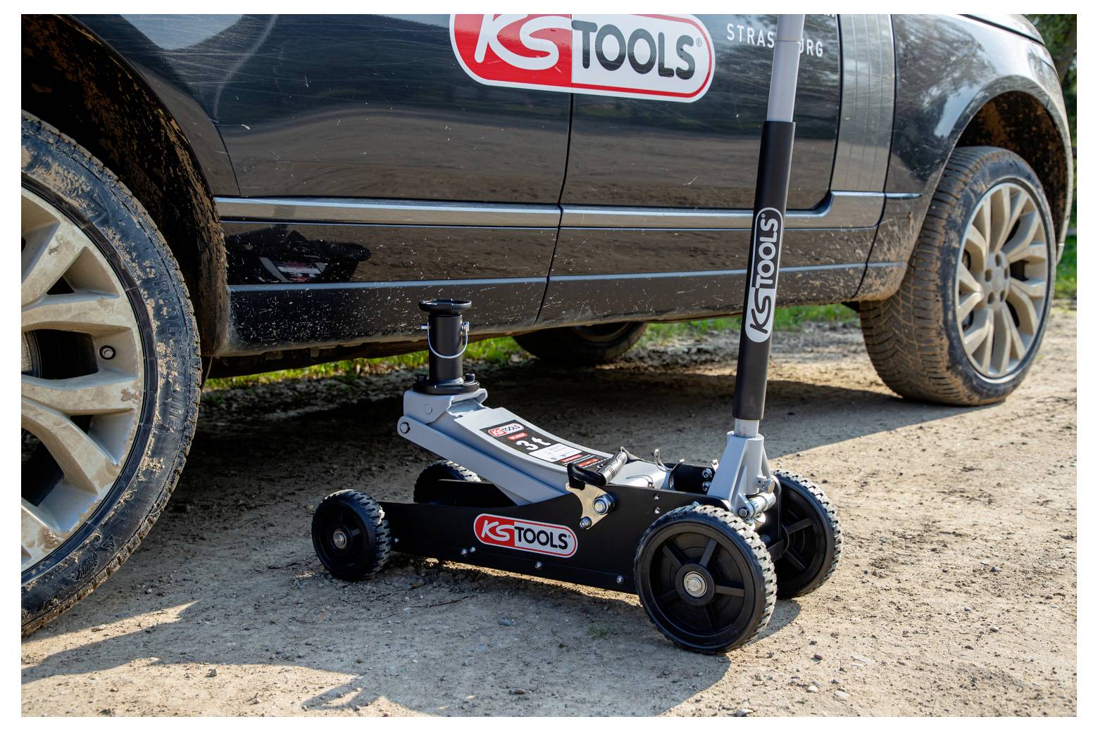 Hydraulischer Flachbodenwagenheber mit KSTools-Branding, der neben einem schwarzen SUV angehoben wird, geparkt auf Schmutz in der Nähe des Vorderrads.