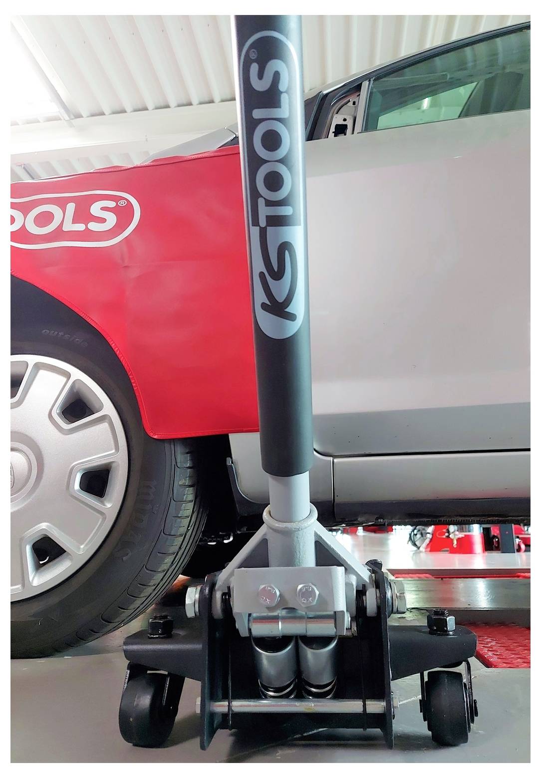 Nahaufnahme eines hydraulischen Wagenhebbers, der ein Auto neben einem anderen Fahrzeug hebt und den Griff des Hebbers, die Räder und den Hebearm unter dem Auto zeigt.