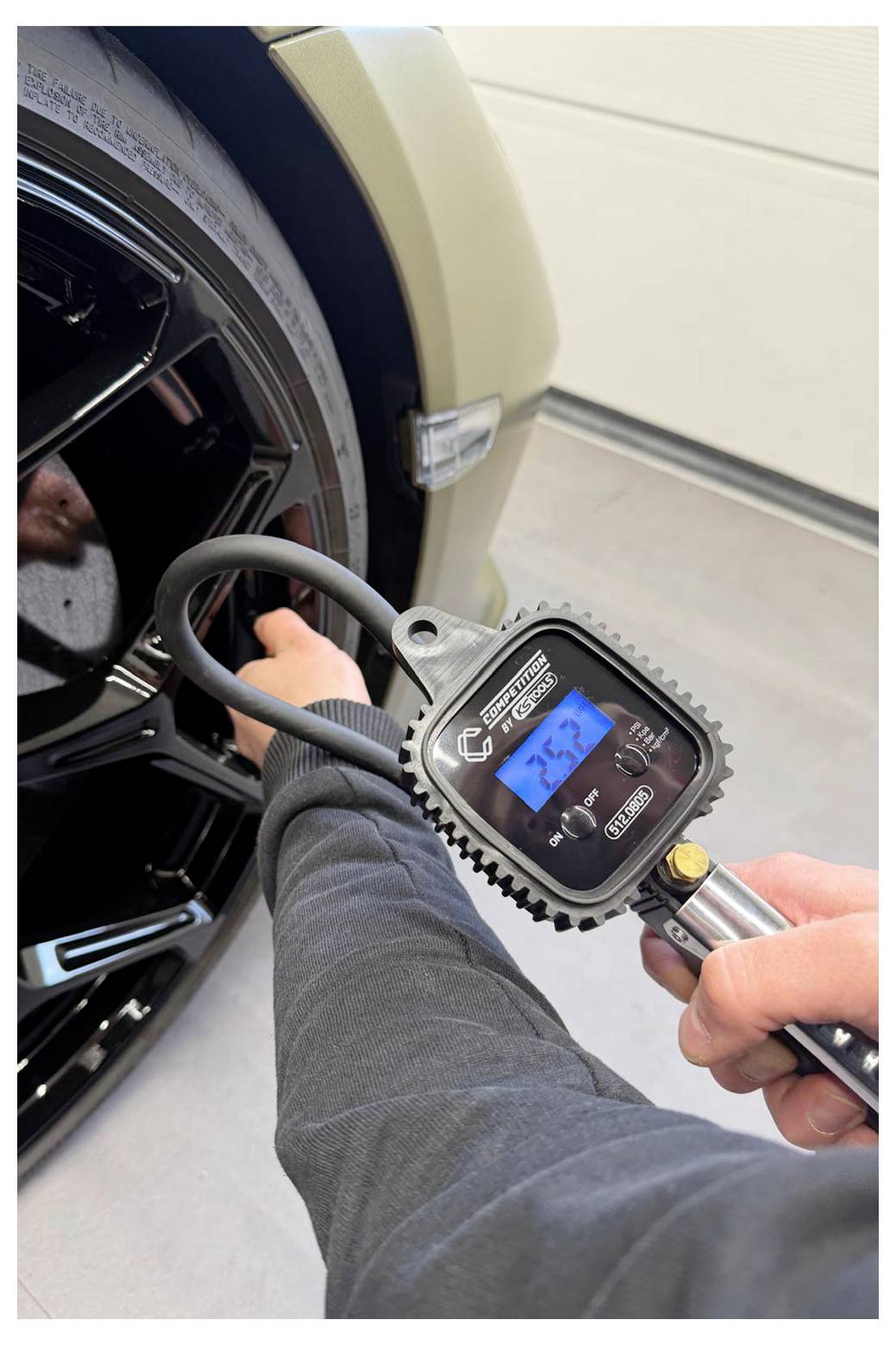 Eine Person bläst einen Autoreifen mit einem digitalen Druckmesser und Luftschlauch in einer Garage auf und zeigt das Reifenventil an einem Fahrzeugrad.