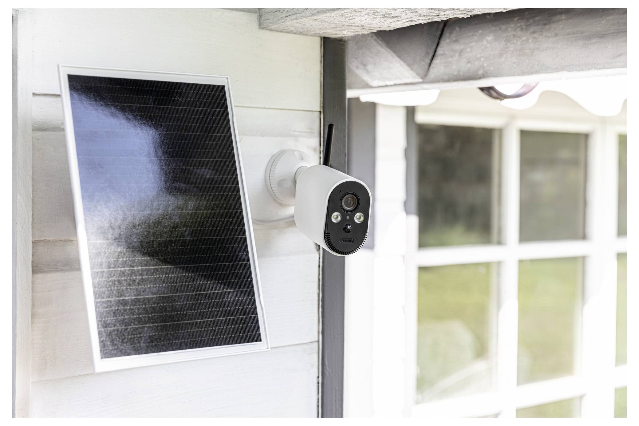 Solar-betriebene Sicherheitskamera an der Außenseite eines Hauses neben einem Solarpanel montiert, ausgerichtet auf den Hof oder die Haustür.