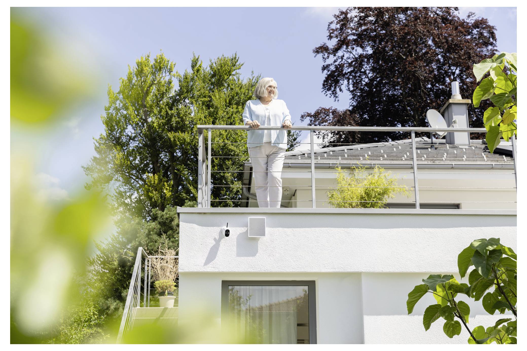 Eine ältere Frau steht auf einer modernen Dachterrasse eines Hauses, lehnt sich an das Geländer und blickt an einem sonnigen Tag nach außen.
