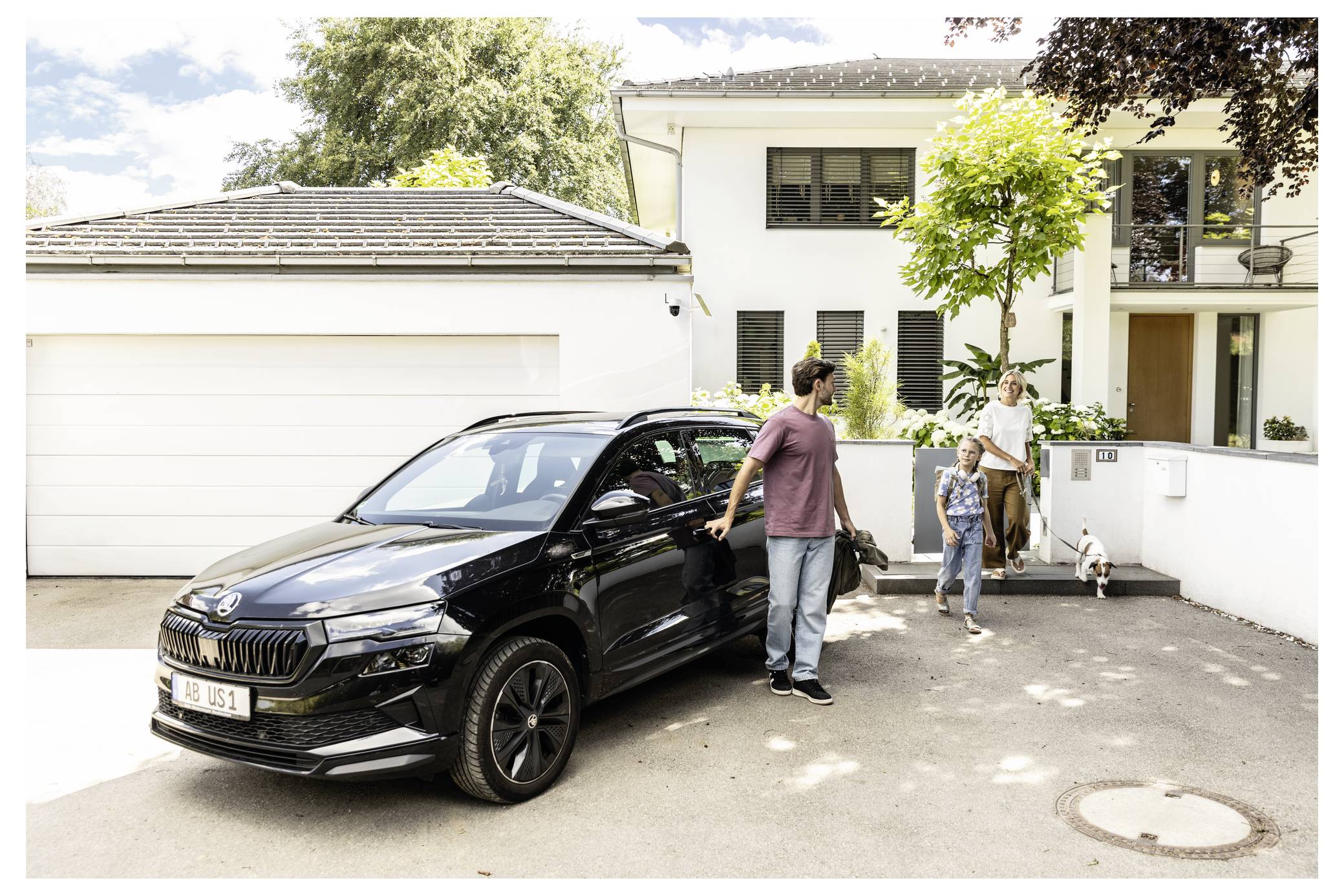 Eine Familie steht neben einem schwarzen SUV in einer Einfahrt vor einem modernen weißen Haus, mit einem Kind und einem Hund in der Nähe des Eingangs.