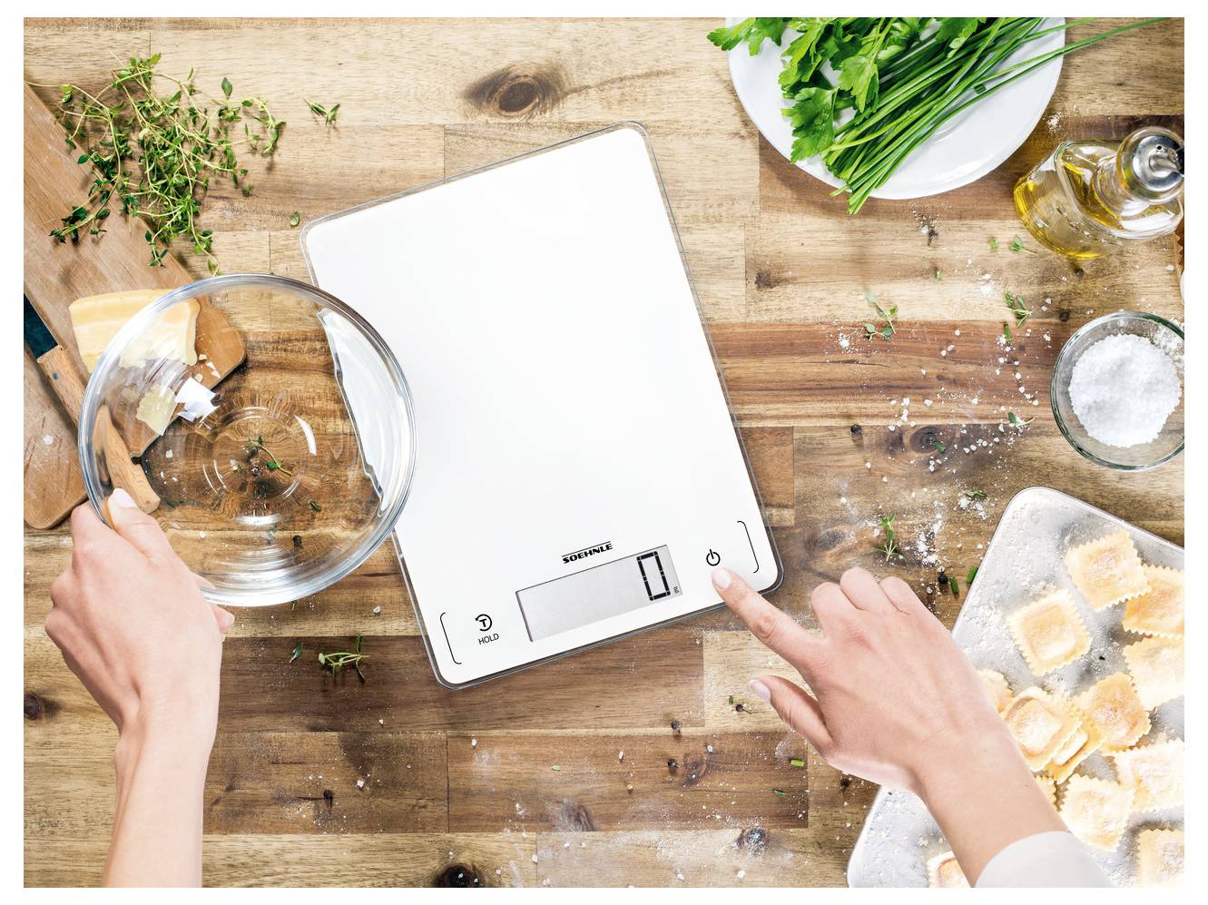 Hände bereiten sich darauf vor, eine digitale Küchenwaage auf einem Holztisch zu verwenden, mit Kräutern, Öl, Salz und Zutaten in der Nähe.