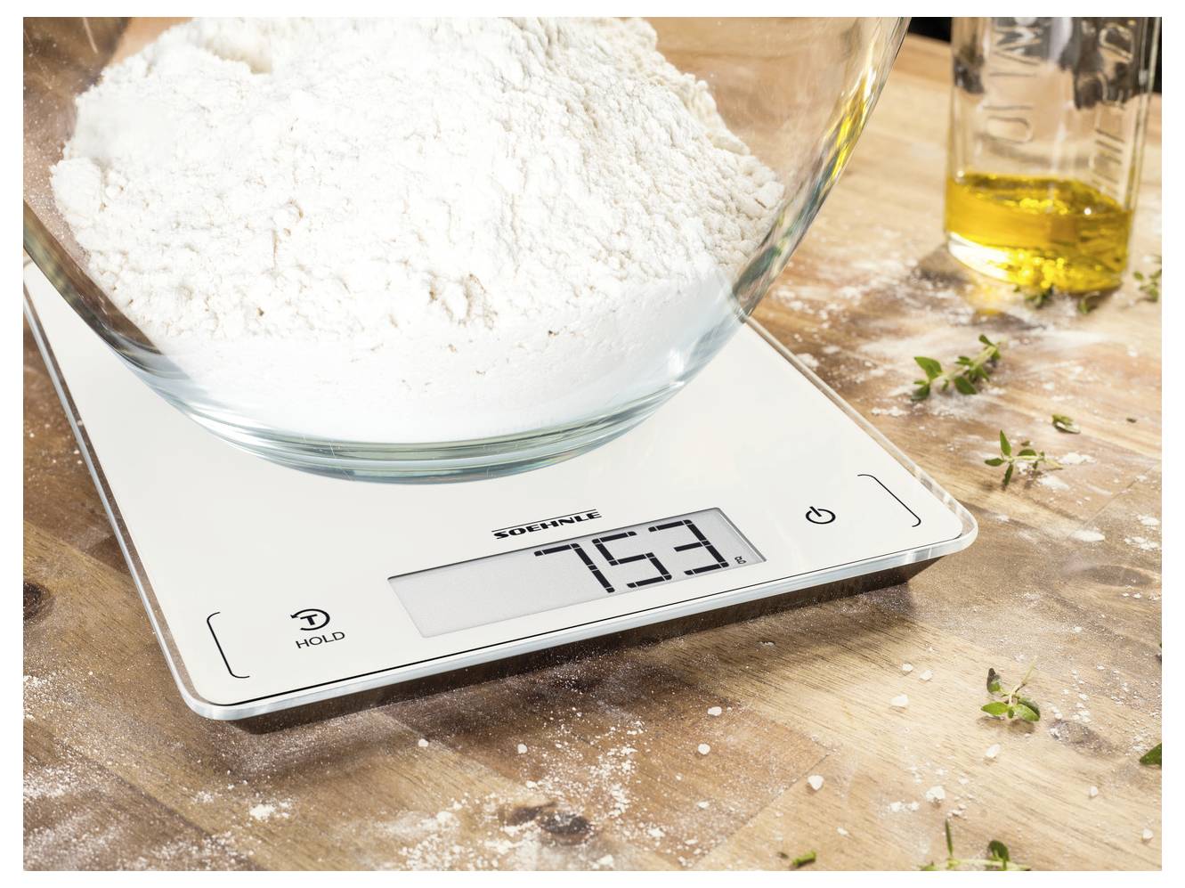 Glasschüssel mit Mehl auf einer digitalen Küchenwaage, die 753 Gramm anzeigt, auf einer bemehlten Holzarbeitsplatte mit Kräutern und Olivenöl in der Nähe.