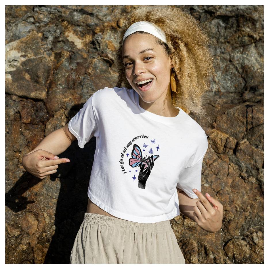 Lächelnde Frau im Freien zeigt auf eine Schmetterlingsgraik auf ihrem weißen T-Shirt mit dem Text „Ich lasse alle meine Sorgen los".