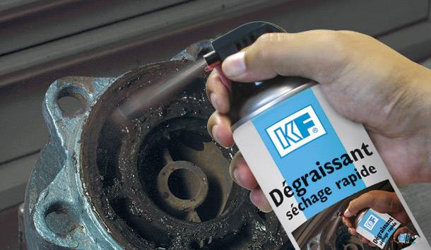 Eine Person besprüht einen rostigen Metallmechanismus mit einem Spray aus einer Dose, beschriftet als 'KF Dégraissant séchage rapide'.