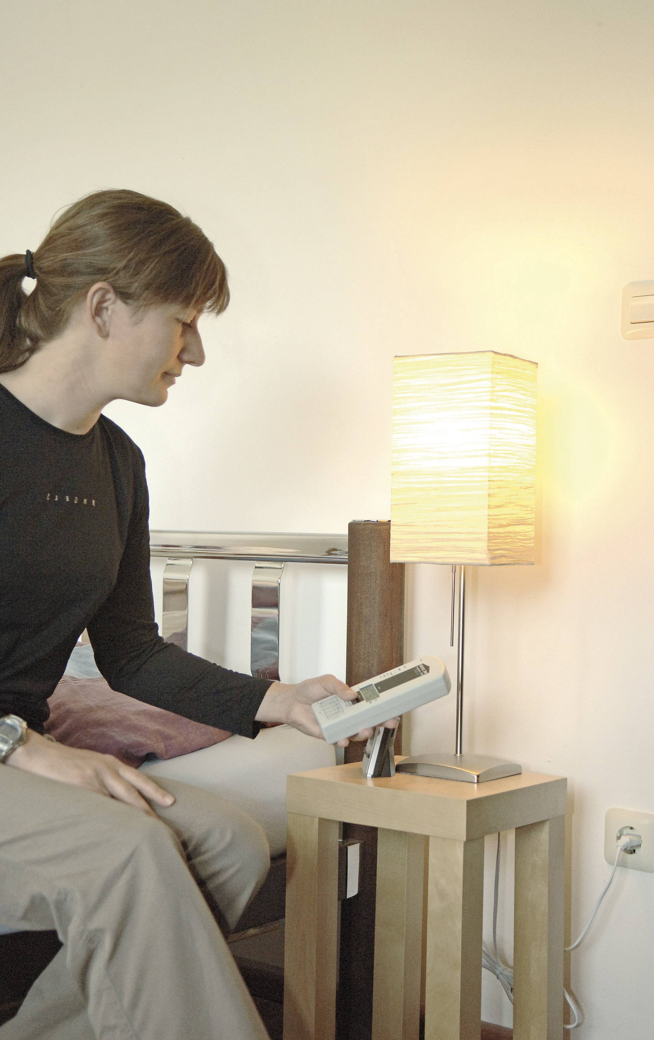 Person sitzt auf Bett, hält Fernbedienung, richtet sie auf einschaltbare Tischlampe auf Nachttisch. Lampe leuchtet.