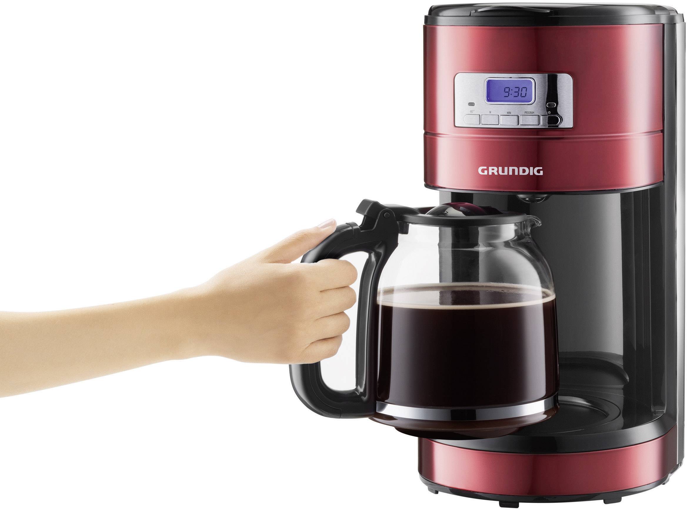 Rote Kaffeemaschine mit Glaskanne, gefüllt mit Kaffee. Eine Hand hält den Kannenhenkel. Digitalanzeige zeigt Uhrzeit 9:30.