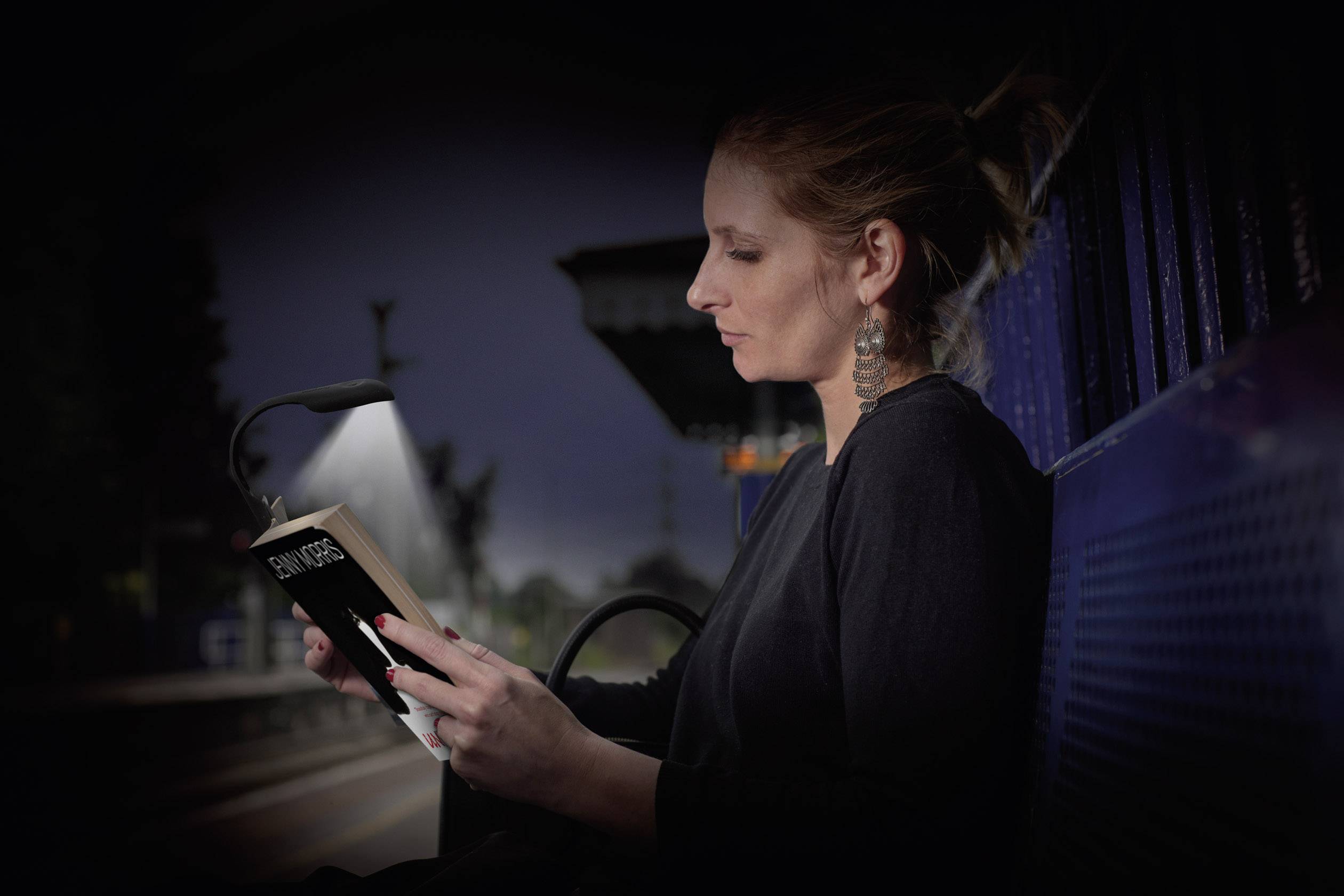 Eine Person liest bei Nacht ein Buch auf einem Bahnsteig, beleuchtet von einer kleinen Lampe, auf einer Bank sitzend.