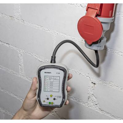 Eine Hand hält ein Gerät zur Überprüfung elektrischer Anschlüsse. Es ist mit einer roten Steckdose an einer Backsteinwand verbunden.