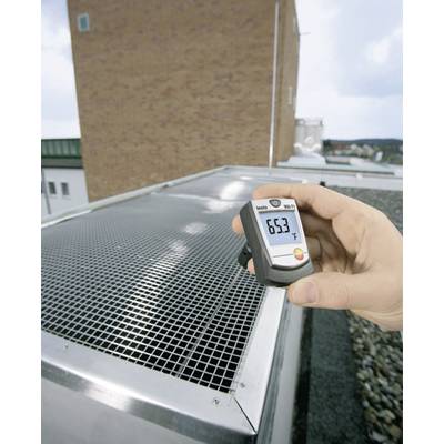 Eine Hand hält ein digitales Thermometer, das 63 Grad Fahrenheit anzeigt, über einem Außendachgitter eines Gebäudes. Hintergrund zeigt Himmel und Gebäude.