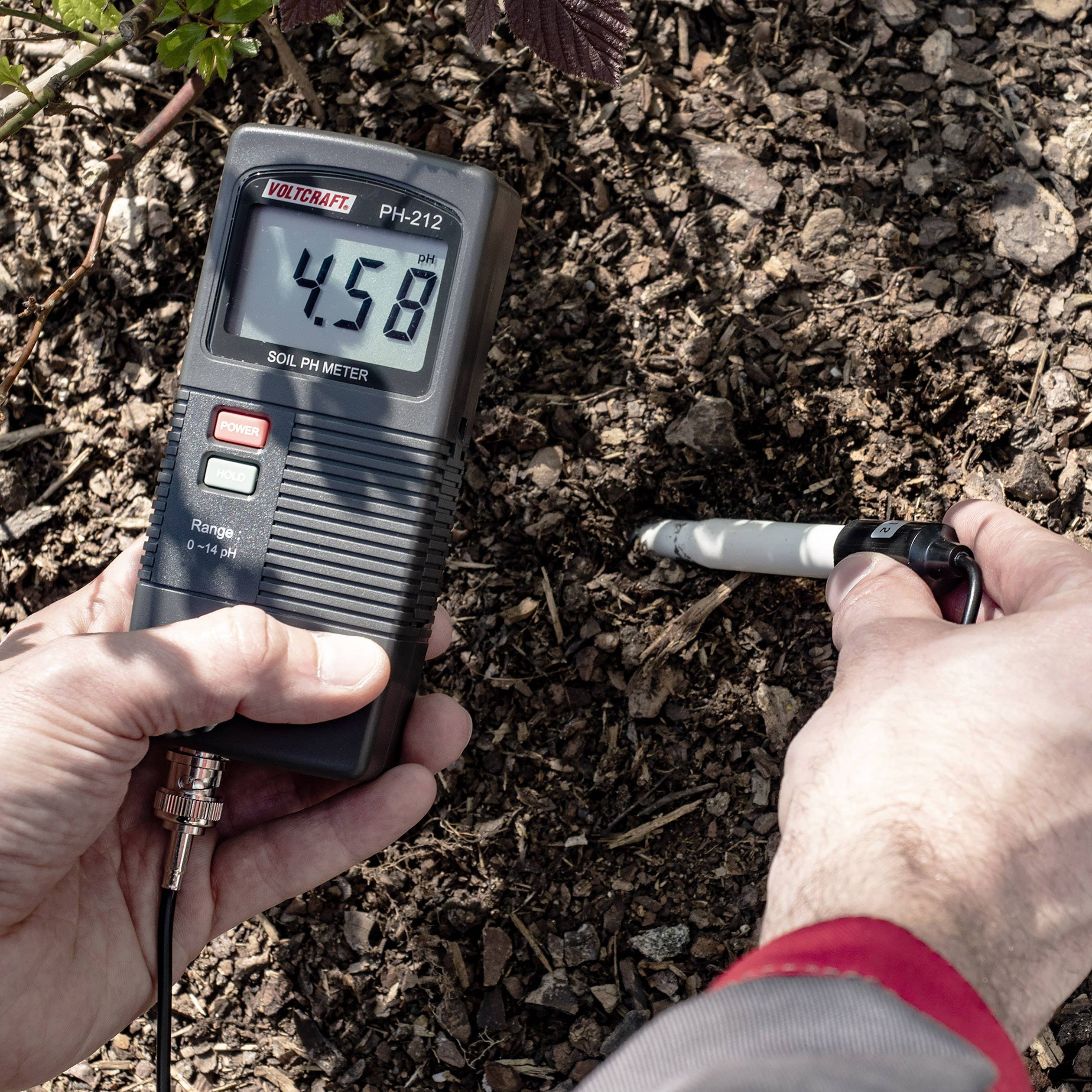 Eine Person misst mit einem digitalen pH-Meter den pH-Wert des Bodens im Garten. Anzeige zeigt '4.58', niedriger pH-Wert.