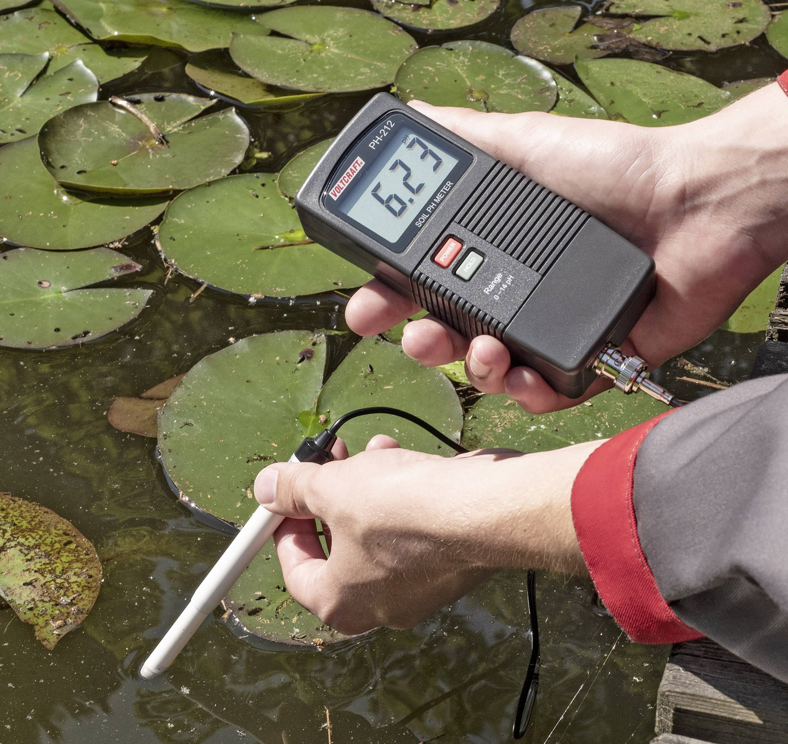 Eine Person hält ein digitales Messgerät über einem Teich mit Seerosenblättern. Das Gerät zeigt '6,23' an.