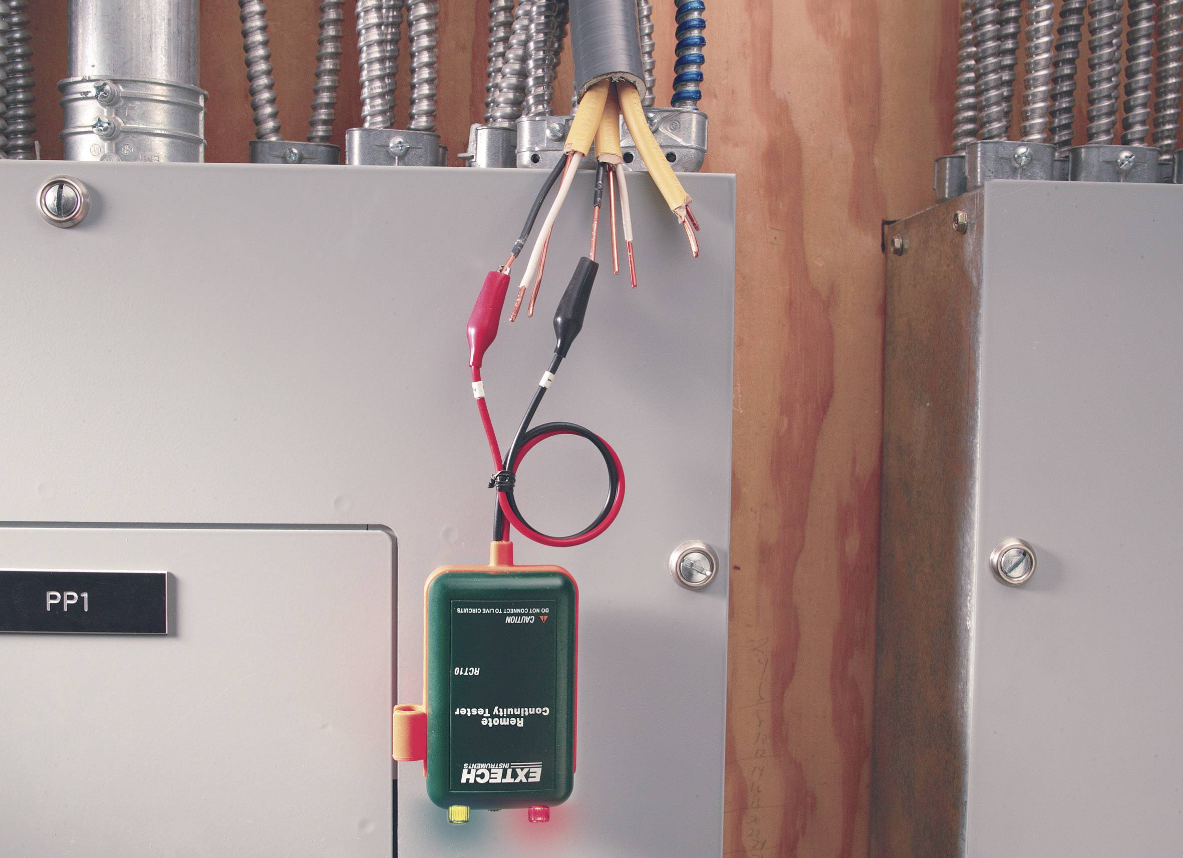 Ein elektrisches Prüfgerät ist an freiliegende Kabel angeschlossen und zeigt Spannungsmessungen an. Es hängt vor einer Schalttafel.