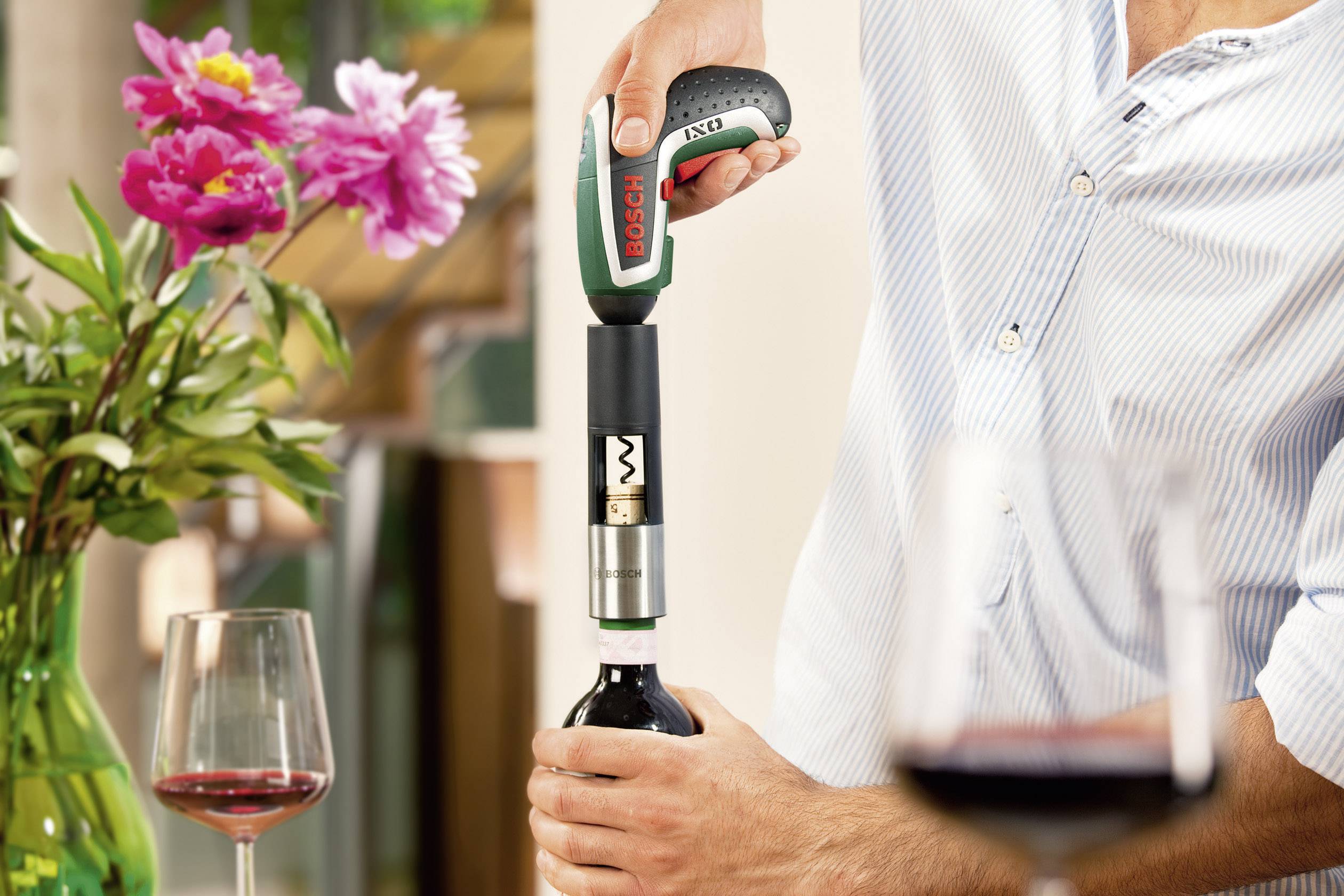 Eine Person öffnet eine Weinflasche mit einem elektrischen Korkenzieher in einer häuslichen Umgebung. Im Hintergrund Blumen und Weingläser.