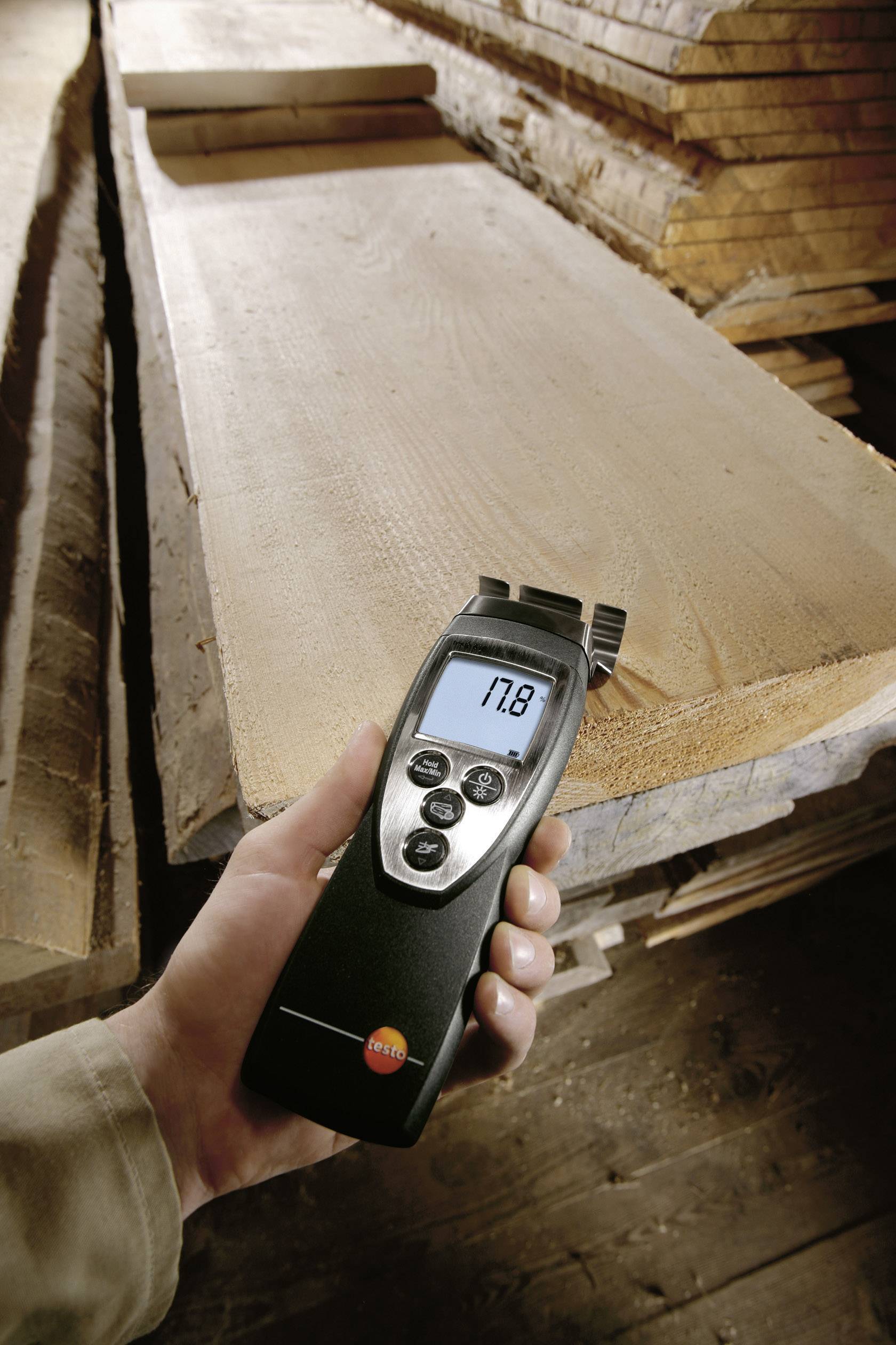 Eine Hand hält ein digitales Feuchtigkeitsmessgerät, das '17,8' anzeigt, über einem Stapel von Holzplatten in einer Werkstatt.