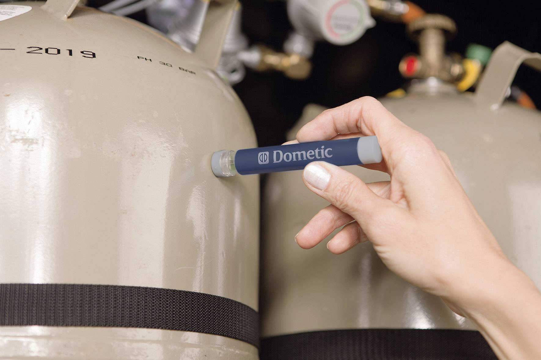 Eine Hand hält ein Thermometer an einen grauen Gastank, um den Füllstand zu prüfen. Der Tank zeigt die Aufschrift '2019'.