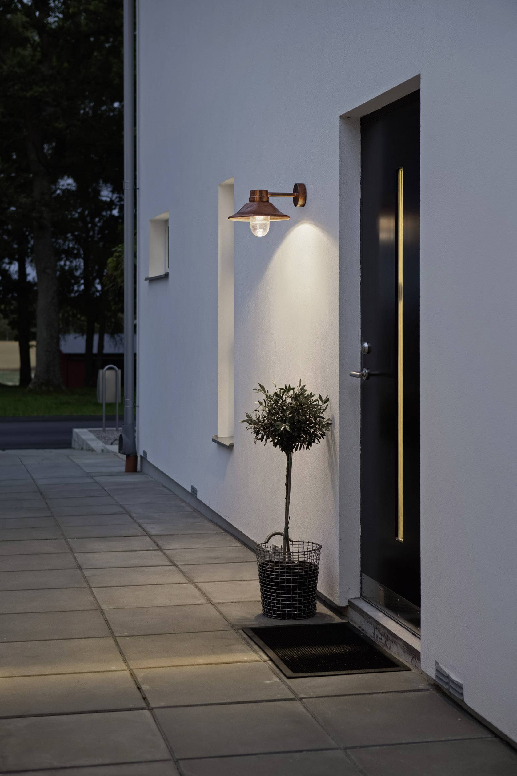 Moderne Außenwandlampe beleuchtet den Hauseingang neben einem kleinen Baum in einem Topf auf einer Fliese.