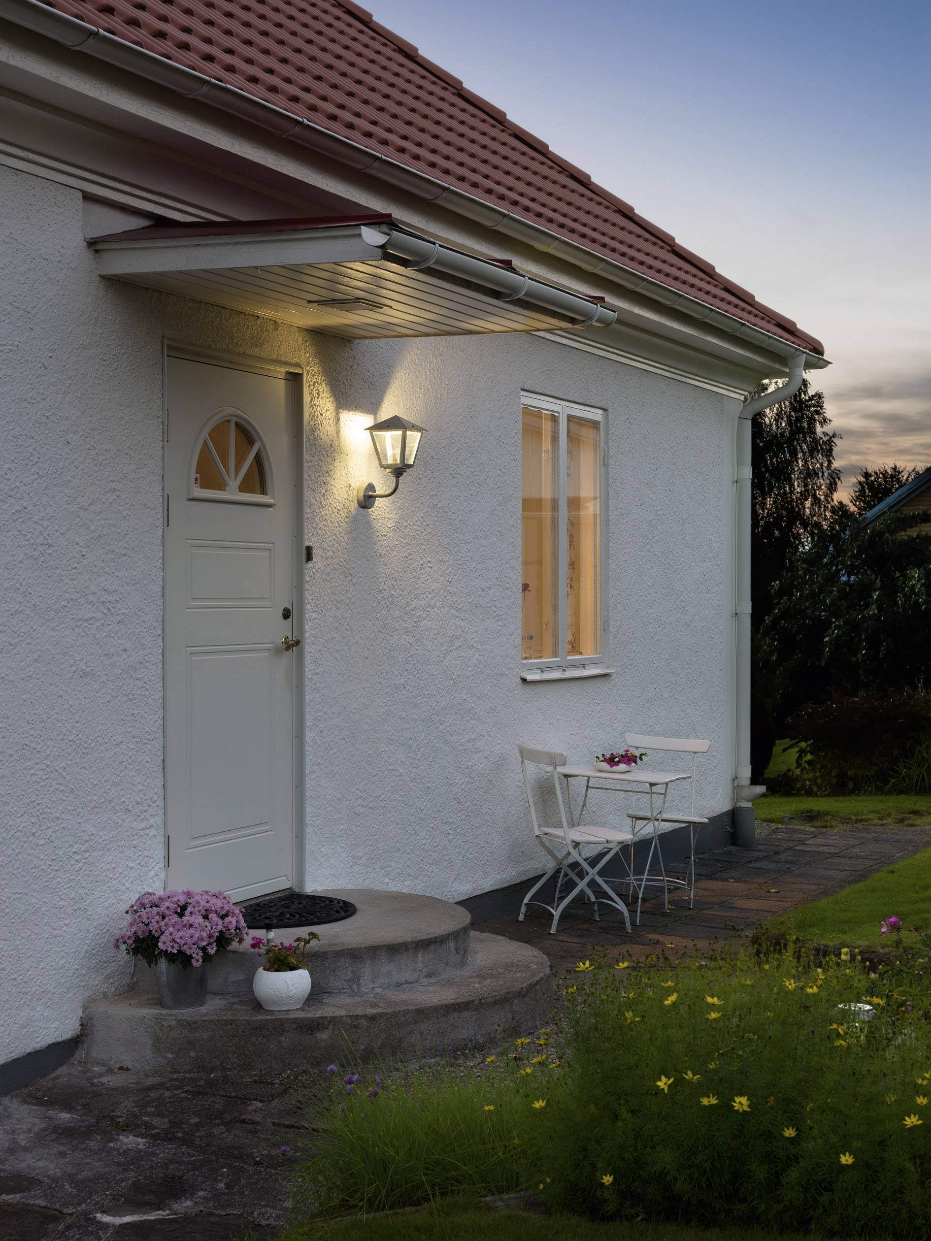 Eine weiße Hausfassade bei Dämmerung mit beleuchteter Haustür. Ein kleiner Tisch und zwei Stühle stehen davor, umgeben von Blumen.